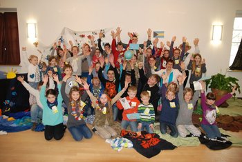 Kinder und Jugend Eine große Schar fröhlicher Kinder sitzt und steht in einem Pfarrsaal und hält gemeinsam einen ökumenischen Kinderbibeltag
